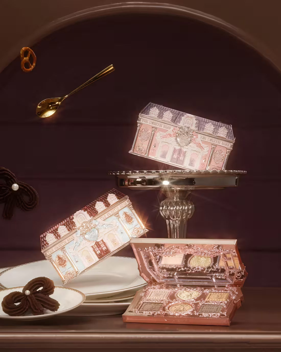 Three variants of Flowerknows Sweetie Bear eyeshadow palettes arranged on a decorative plate against a rich brown background. One palette sits open at the bottom revealing the matte and shimmer shades, while the other two stand vertically erect, showcasing the intricate vintage-style neoclassical architecture packaging.