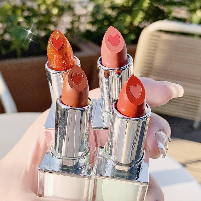 Four lipsticks with heart-shaped caps on a reflective surface outdoors.