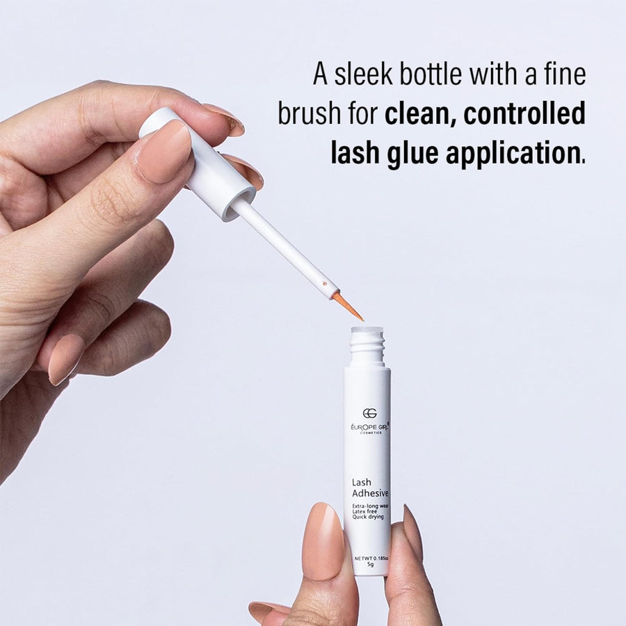 Hand holding a bottle of lash adhesive with a fine brush, against a light gray background.