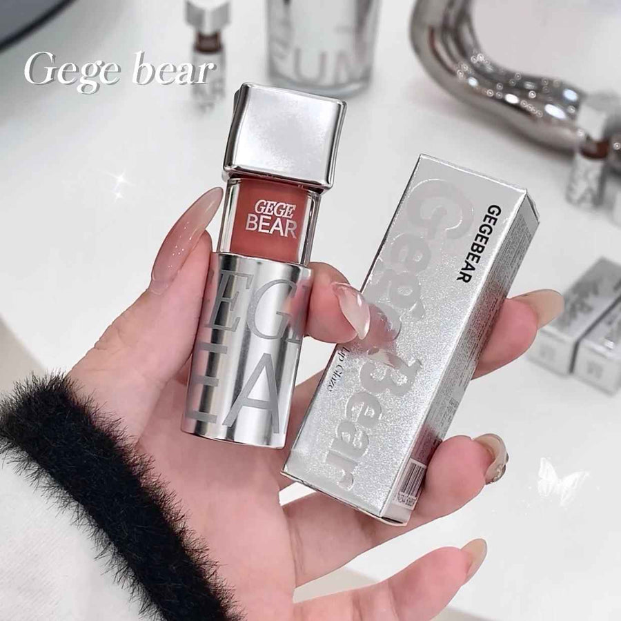 Hand holding a silver lip gloss with 'Gege Bear' branding against a blurred bathroom background.
