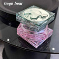 Pack of three Gege Bear cushion matte liquid foundations displayed on a black glass table, featuring light green and pink packaging colors with a clean product showcase setup.