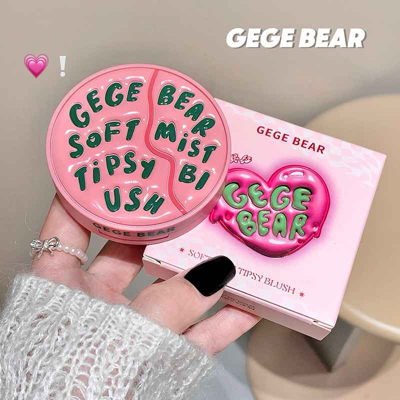 Pink 'GEGE BEAR' makeup blush with green text held by a hand against a neutral background.