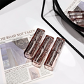 Three transparent lipstick on a newspaper with a mirror and pink items in the background.