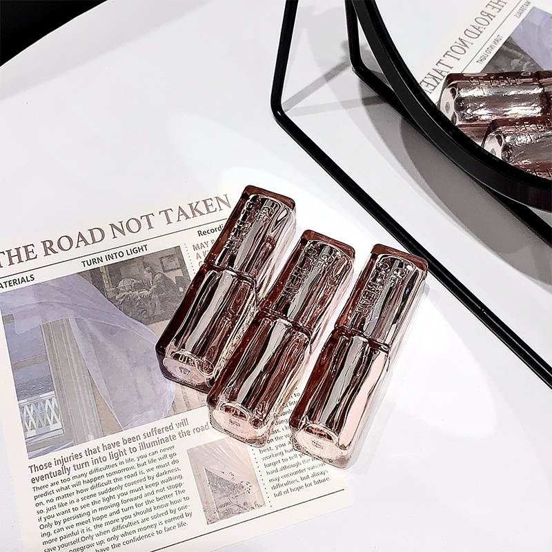 Three transparent lipstick on a newspaper with a mirror and pink items in the background.