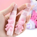 Two pink bear-shaped lip glaze containers with 'GEGE BEAR' text held in a hand against a blurred background.