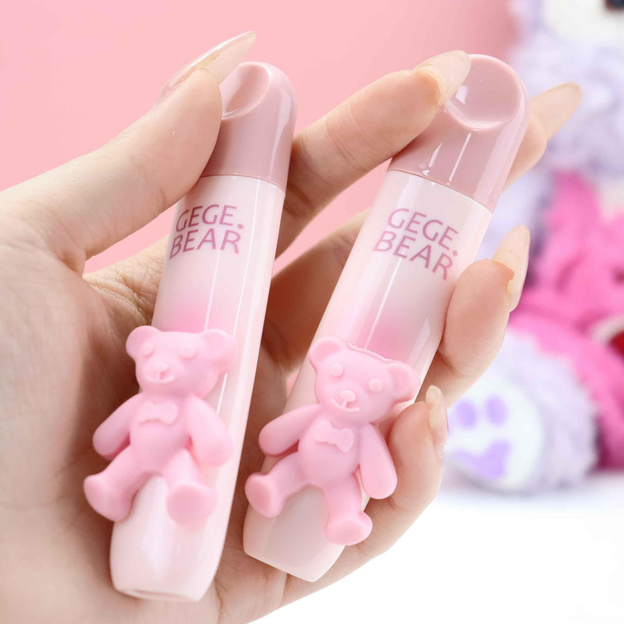 Two pink bear-shaped lip glaze containers with 'GEGE BEAR' text held in a hand against a blurred background.