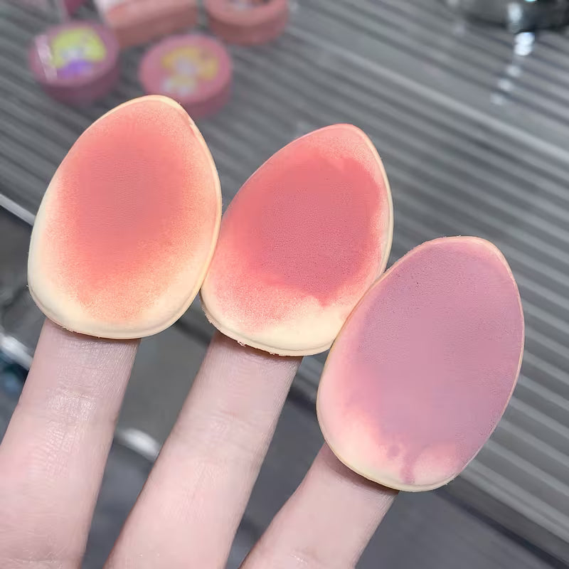 Three pink makeup sponges held between fingers with a blurred background