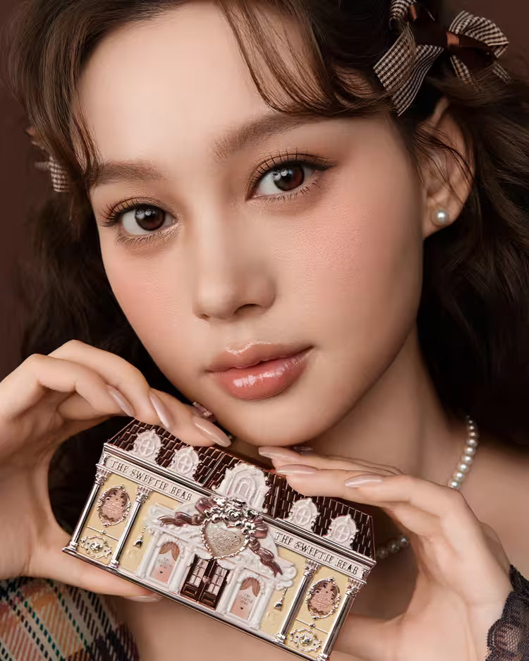 A close-up of a girl holding Flowerknows Sweetie Bear eyeshadow palette with both hands, showcasing swatches of the romantic nude pink and warm neutral matte shades pigmented on her skin.