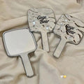 Two hand-held mirrors with 'Gege Bear' branding on a beige fabric background.