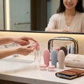 a girls in a bathroom using a travel bottle in a pink color to pump out the clear lotion from the bottle around her there are more bottles and kit in her cosmetic bag 